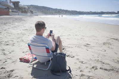 Rear view of man using smart phone while sitting on chair at beach