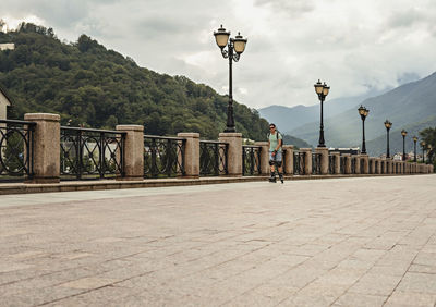 Young man in protective equipment  riding on roller skates along embankment against mountains 