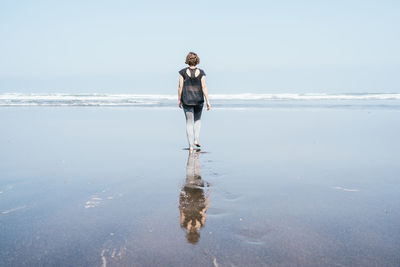 Full body back view of unrecognizable barefooted female in sportswear walking on wet sand towards waving sea