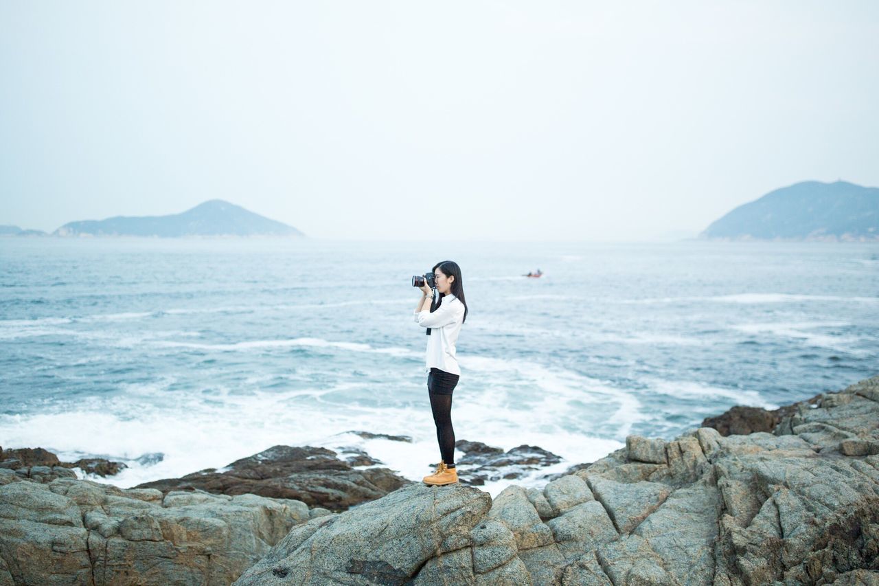 full length, water, sea, lifestyles, standing, leisure activity, rock - object, scenics, beauty in nature, tranquil scene, horizon over water, tranquility, rear view, nature, clear sky, shore, casual clothing, mountain