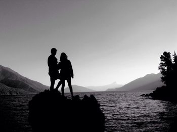 Silhouette couple standing on rock amidst river against clear sky