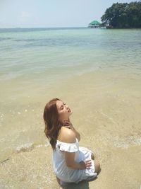 Young woman sitting at beach against sky