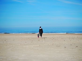 Rear view full length of mother with daughter walking on sand at beach against blue sky