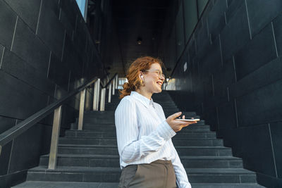 Happy businesswoman with smart phone talking through wireless in-ear headphones