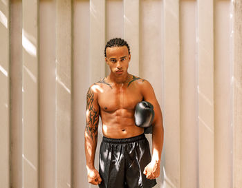 Portrait of shirtless man with boxing glove standing against wall