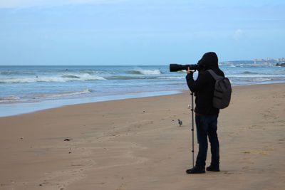 Side view of man photographing while standing at beach against sky