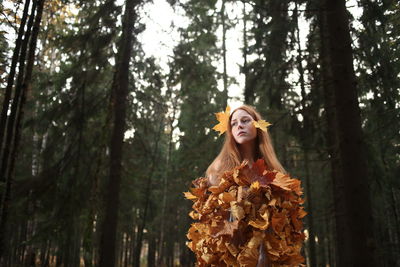 Thoughtful young woman covered with leaves standing at forest during autumn