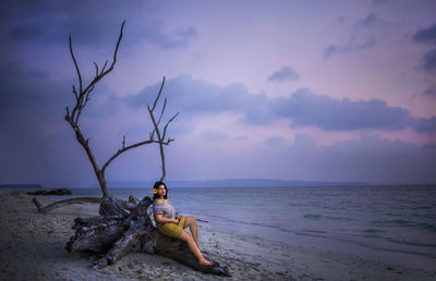 Woman on tree trunk on beach against sky during sunrise