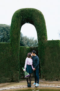 Full length of couple kissing on mouth while standing against hedge in park
