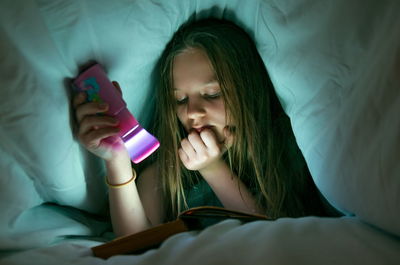 Close-up of girl reading book with illuminated flashlight below blanket on bed