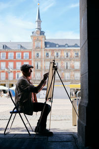 Man painting while sitting against buildings in city