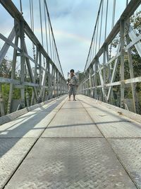 Boy standing on footbridge against sky