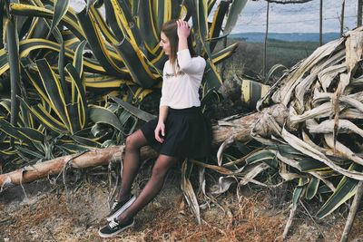 Full length of fashionable young woman with hand in hair sitting on fallen tree