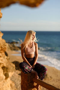 Full length of young woman sitting on wooden log by beach against sea