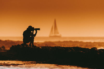 Silhouette man photographing at beach against clear sky during sunset