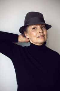 Fashionable older woman in black clothes and a hat stands against a white wall in the office