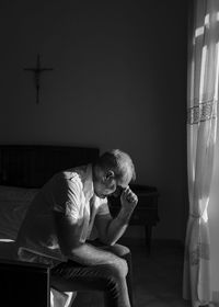 Monochrome of adult man sitting on bed against window with sunlight and shadow