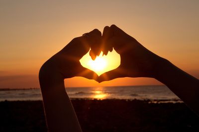 Cropped hands of woman making heart shape at beach against sky during sunset