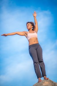 Low angle view of mid adult woman exercising on rock standing against blue sky