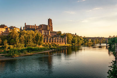 Bridge over river by buildings against sky at sunset