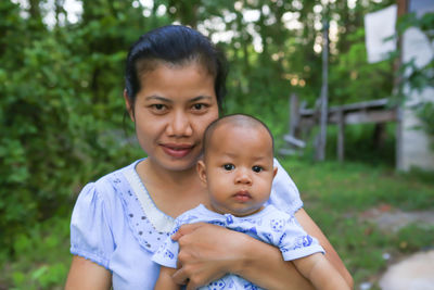 Portrait of woman carrying toddler son while standing against trees