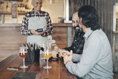 Female bartender with credit card reader talking to male customers at table in bar