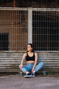 Full length of young woman sitting on skateboard against fence