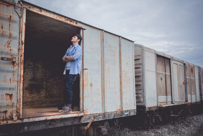Sad young man standing in abandoned freight train against sky