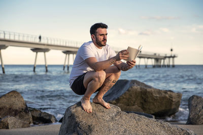 Man reading book on rock at beach against sky