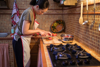 Side view of ethnic female in apron cutting ripe tomatoes on chopping board while cooking lunch in kitchen at home