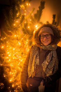 Portrait of smiling woman standing by illuminated christmas tree during winter