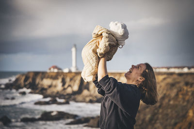 A woman is holding a baby near a lighthouse on the pacific coast