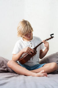 Young boy tuning ukulele at home. blond haired boy sitting on couch playing acoustic guitar.