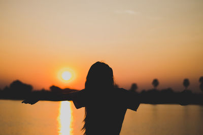 Rear view of silhouette carefree woman with arms outstretched standing by lake during sunset