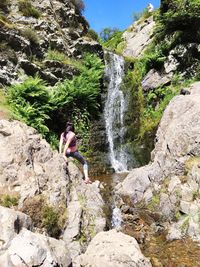 Rear view of woman standing on rock formation by waterfall on sunny day