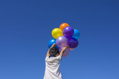 Low angle rear view of boy holding multi colored balloons against clear blue sky