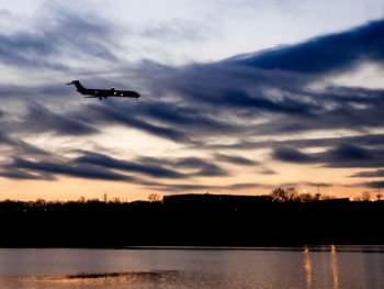 Silhouette airplane flying over lake against sky during sunset