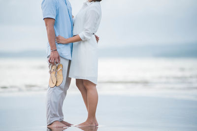 Low section of couple standing at beach against sky