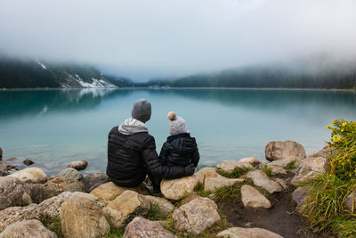 Father and son sitting at lakeshore against sky