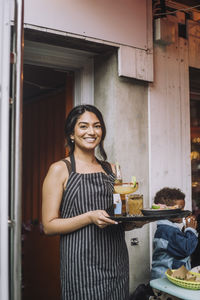 Portrait of smiling waitress wearing apron holding tray of food and drinks at bar