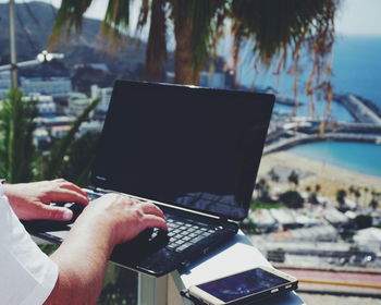 Cropped image of person using laptop by smart phone on sunny day