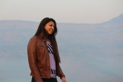Smiling young woman wearing jacket standing against mountain