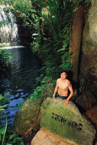 Full length of shirtless man sitting on rock in forest