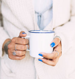 Close view of the hands of a young woman with blue nails and finger rings holding a white enameled 