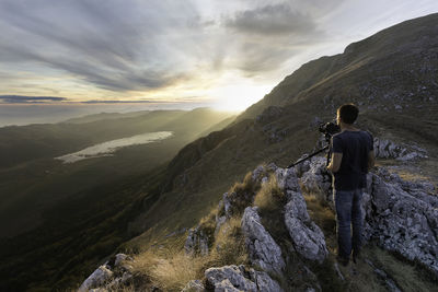 Rear view of man photographing while standing on rock against mountain and sky