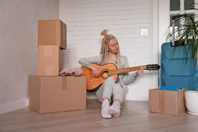 Full length of woman playing guitar while sitting against wall