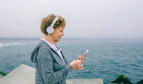 Woman holding smart phone while standing by sea against clear sky