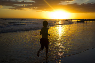 Silhouette boy standing on beach against sky during sunset