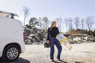 Smiling female electrician carrying ladder and toolbox standing on road by van