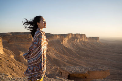 Side view of happy ethnic asian woman in casual clothes standing looking away contemplating the views on cliff in the edge of the world in saudi arabia